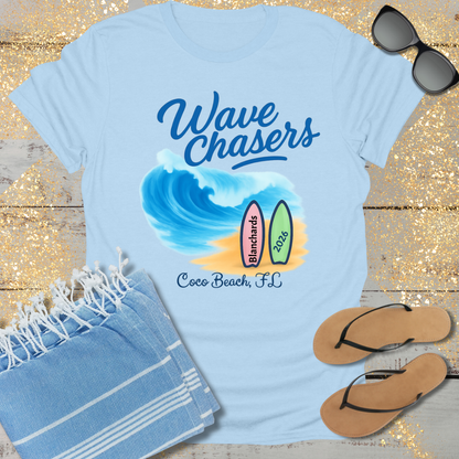 Light blue t-shirt with 'Wave Chasers' design, sunglasses, towel, and flip-flops on a wooden surface.
