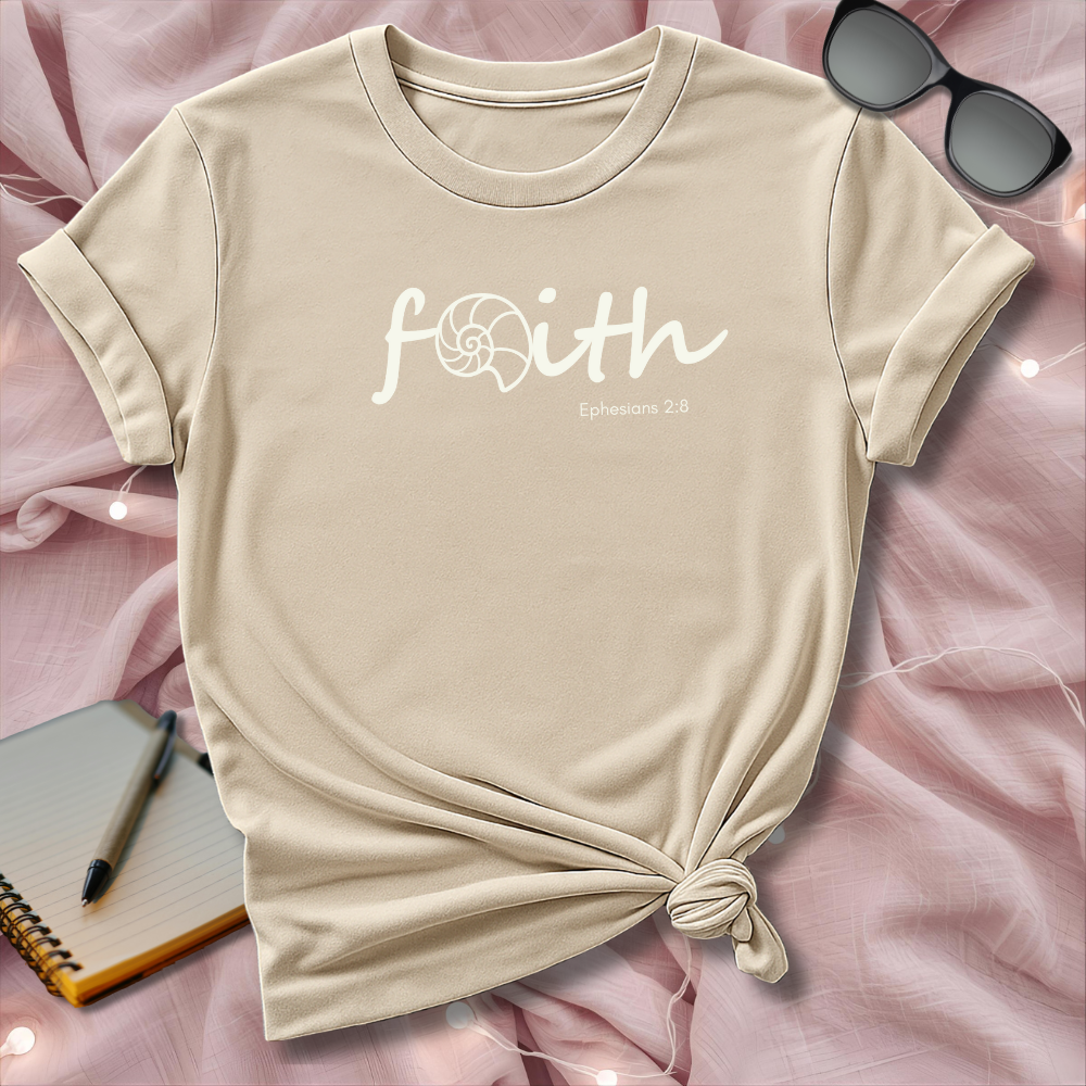Beige t-shirt with 'faith' text on a pink surface with sunglasses and notebook.