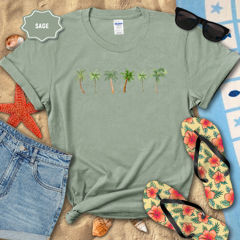 Sage green t-shirt with palm tree design on a sandy surface with beach accessories.