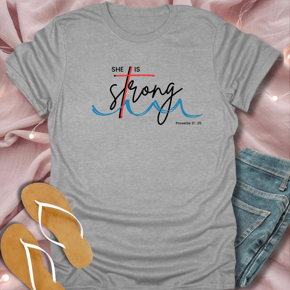 Gray t-shirt with 'She is Strong' text on a pink surface with jeans and sandals.