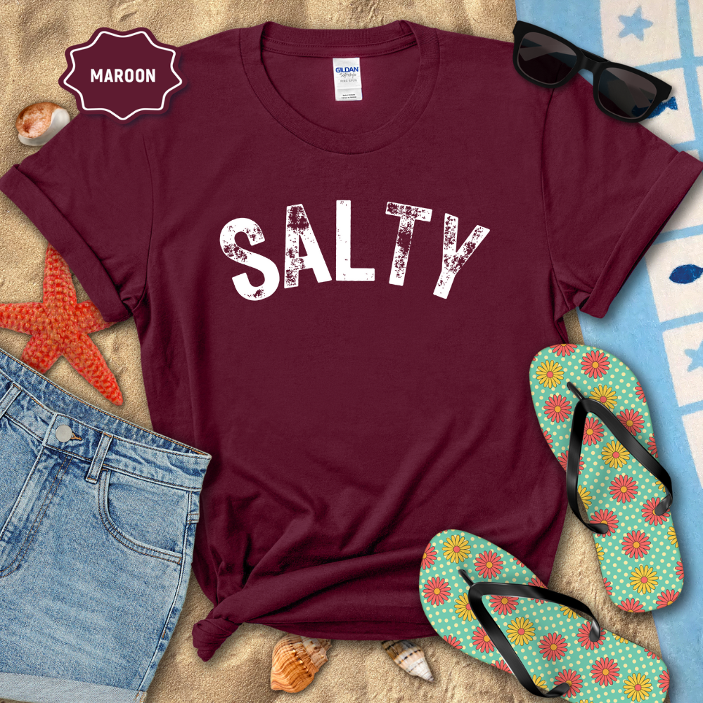 Maroon t-shirt with 'SALTY' text, sunglasses, denim shorts, and flip-flops on a beach-themed background.