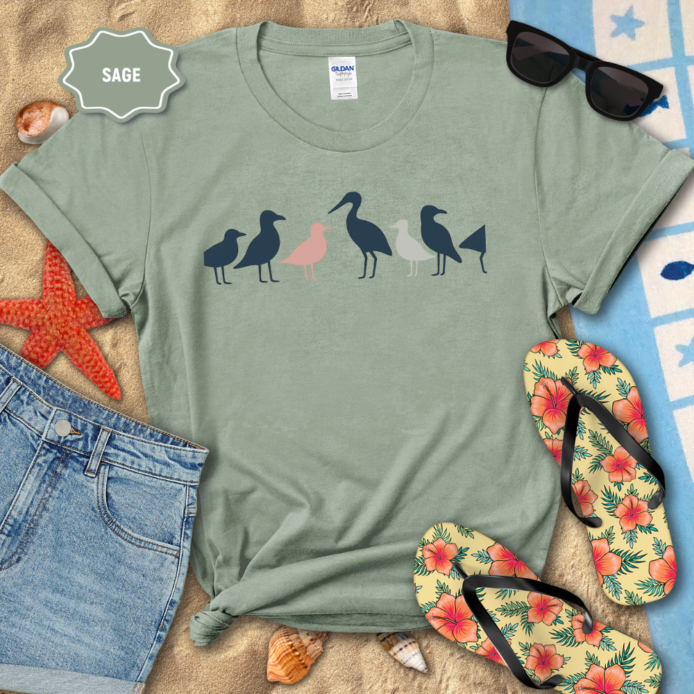 Sage green t-shirt with bird design on a sandy surface with beach accessories.