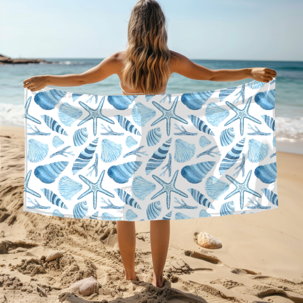 Person holding a beach towel with blue shell and starfish pattern on a sandy beach.