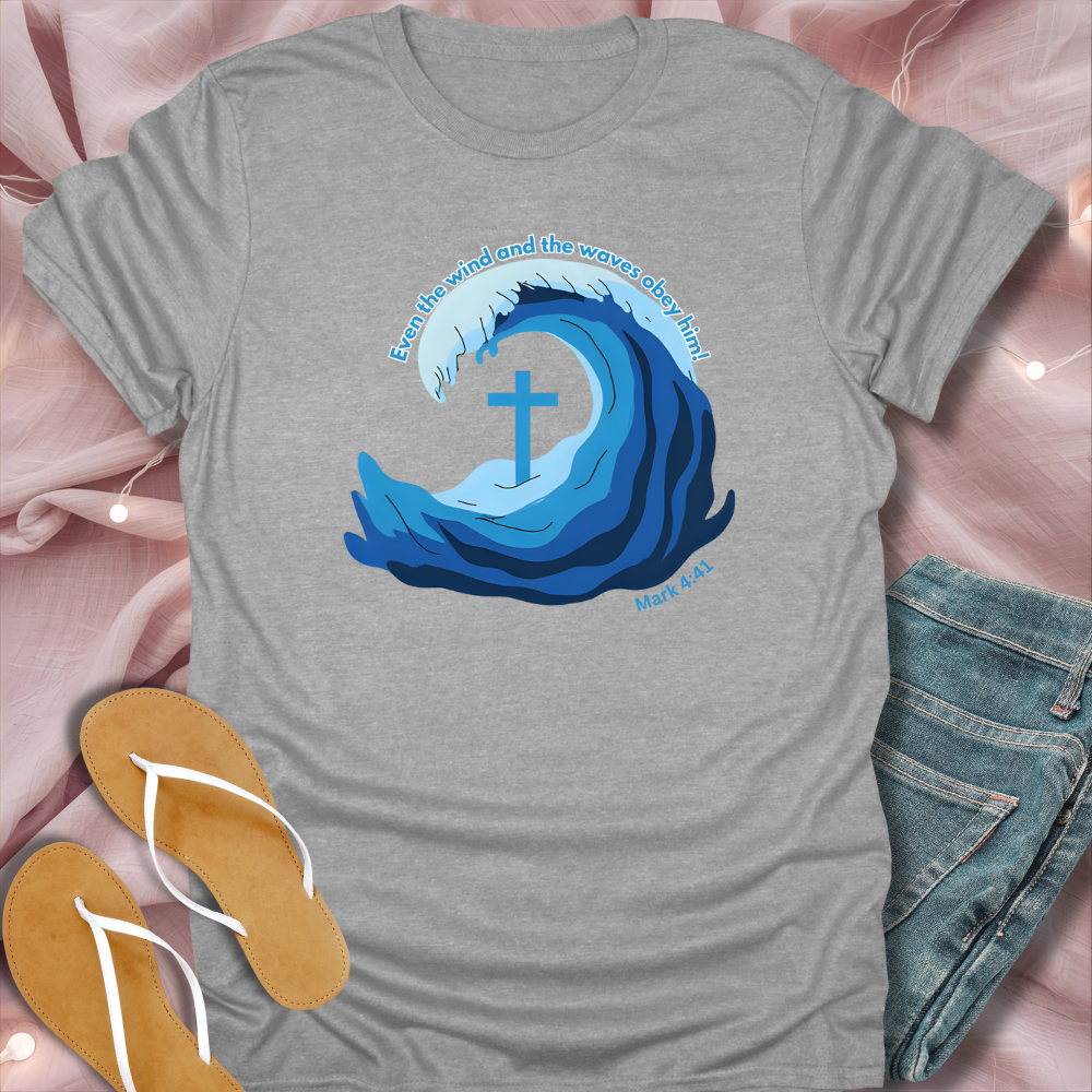 Gray t-shirt with blue wave design and cross, placed on a pink surface with sandals and jeans.
