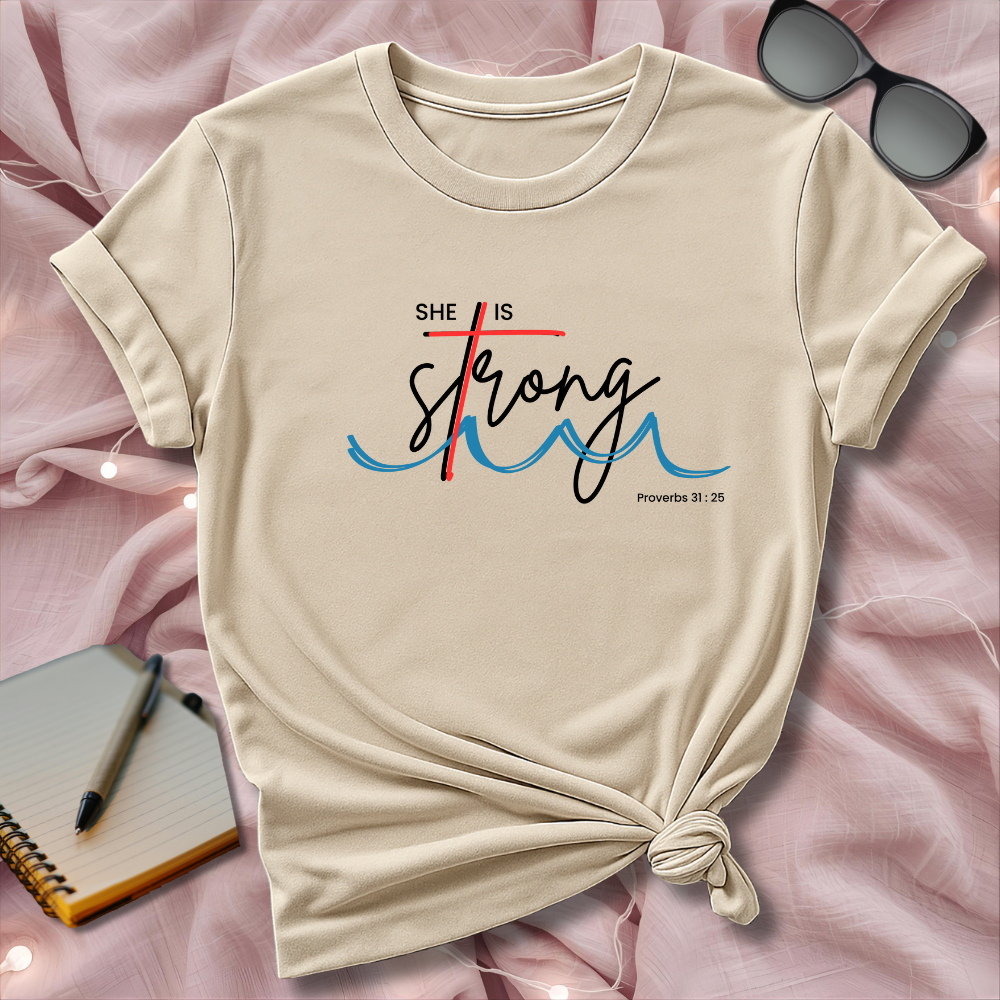Beige t-shirt with 'She is Strong' text on a pink surface with sunglasses and notebook.