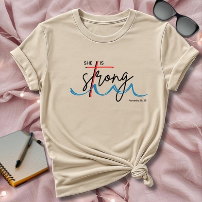 Beige t-shirt with 'She is Strong' text on a pink surface with sunglasses and notebook.
