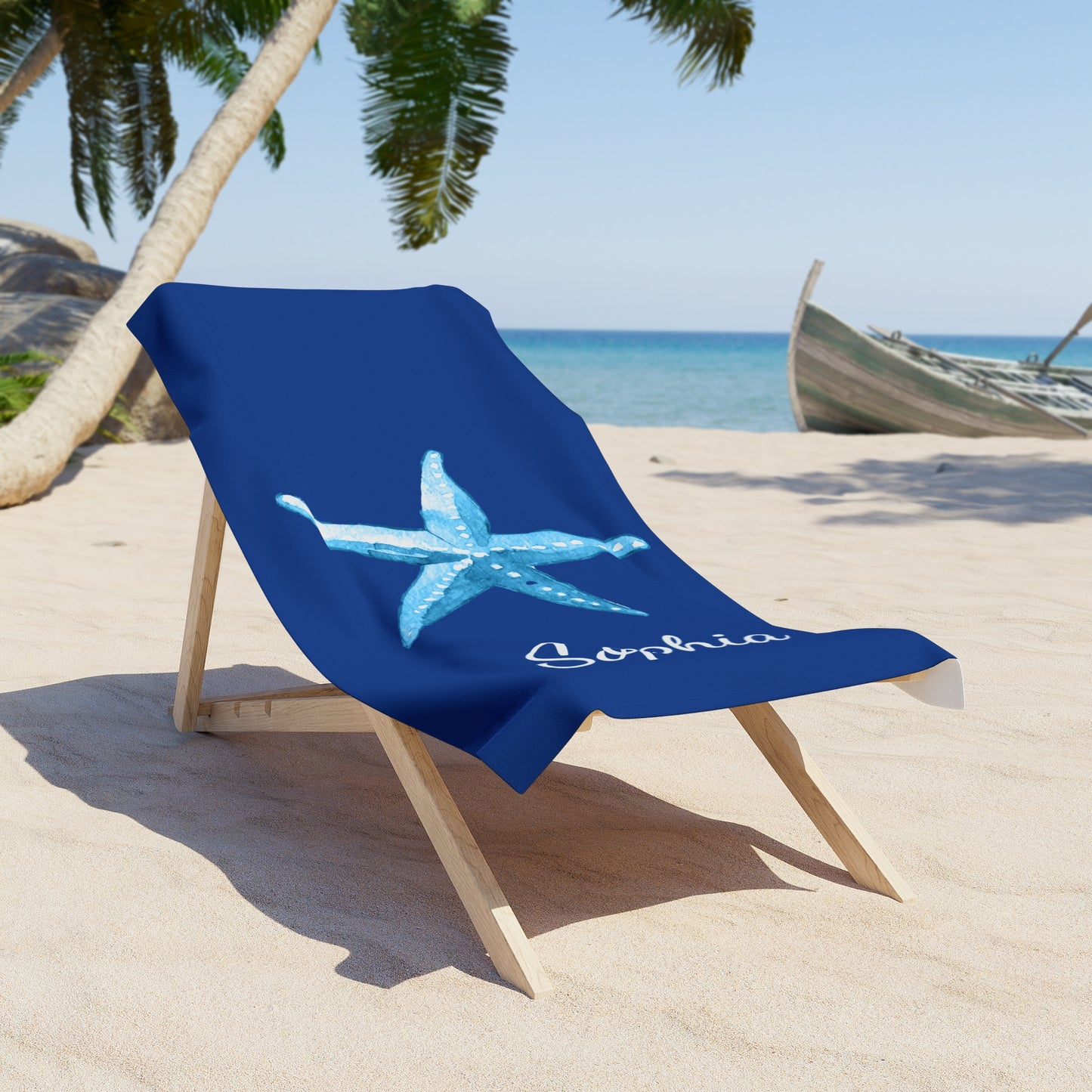 Blue beach towel with a starfish design and 'Sophia' text draped over a wooden chair on a sandy beach.