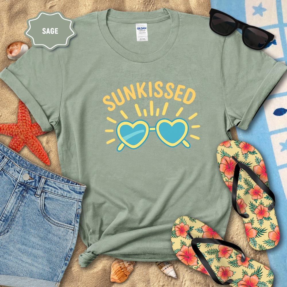 Sage green t-shirt with 'SUNKISSED' design on a sandy surface with beach accessories.