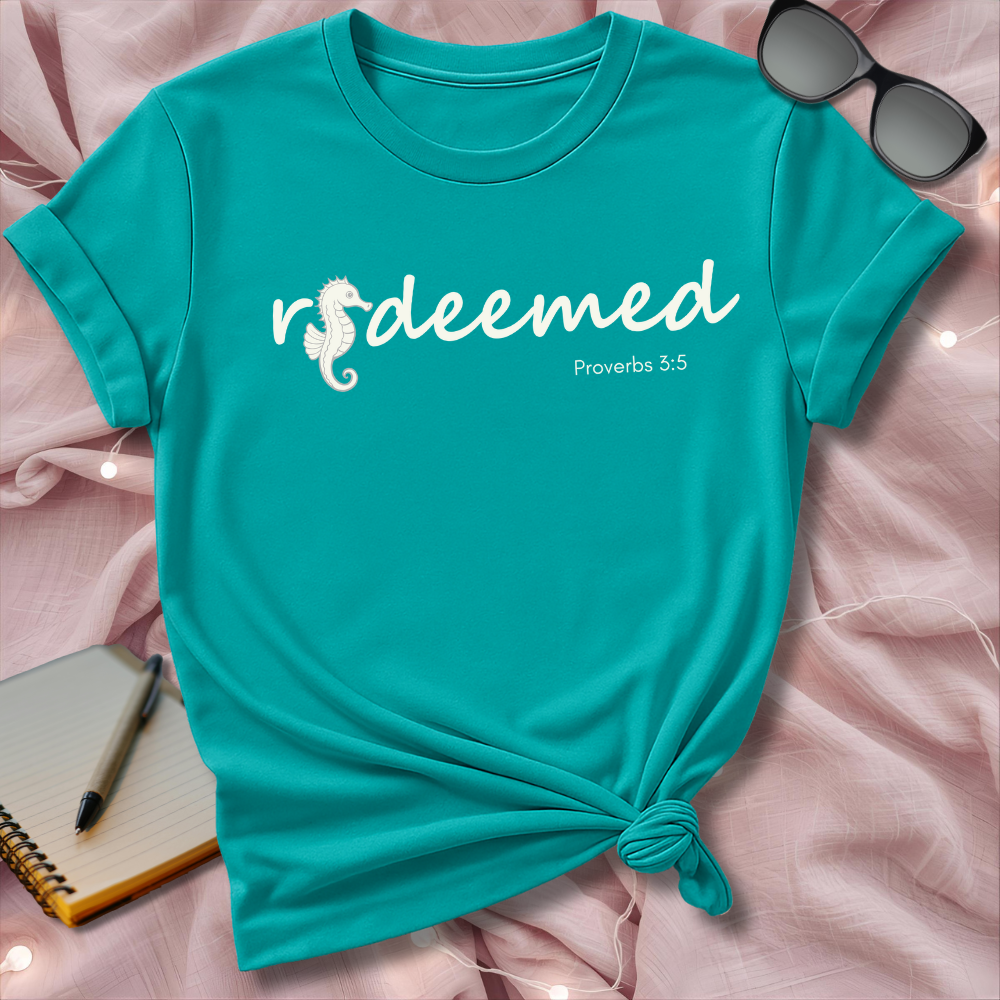 Turquoise t-shirt with 'deemed' text and seahorse graphic on a pink surface with sunglasses and notebook.
