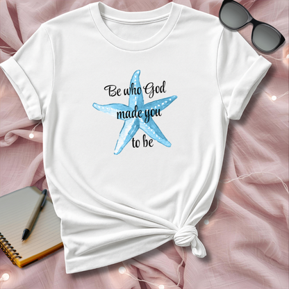 White t-shirt with a blue starfish graphic and text on a pink surface with sunglasses and a notebook.