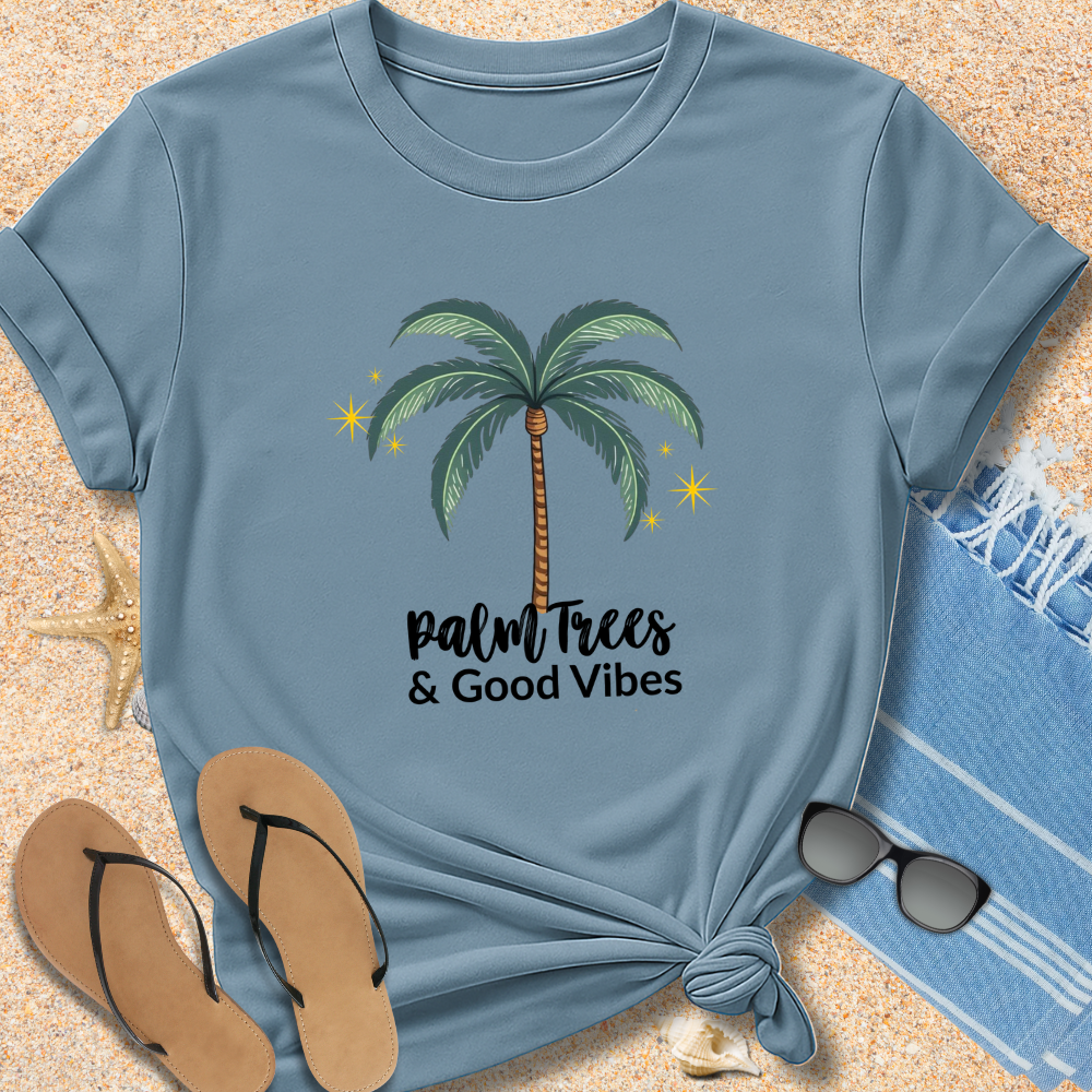 Blue t-shirt with palm tree design and 'Palm Trees & Good Vibes' text on sand with flip-flops and towel.