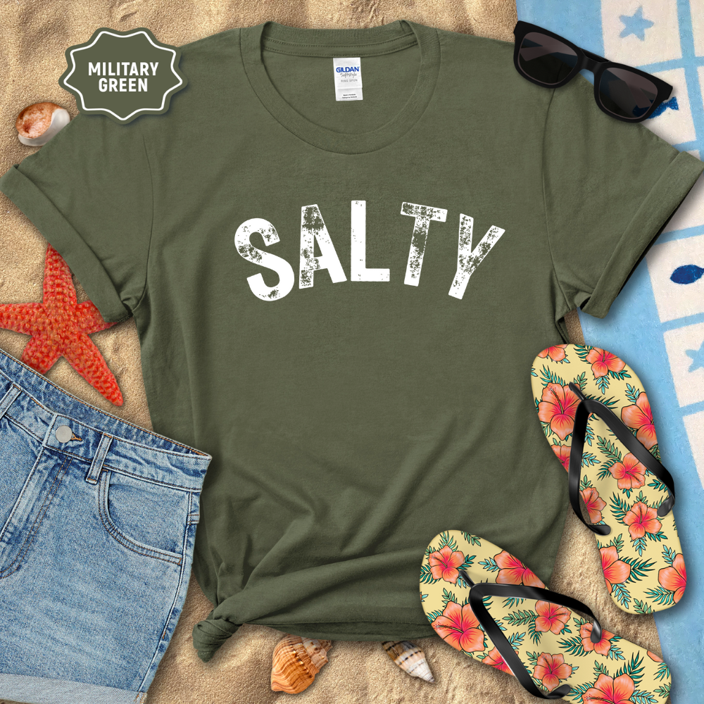 Military green t-shirt with 'SALTY' text, sunglasses, flip-flops, and beach items on a textured surface.
