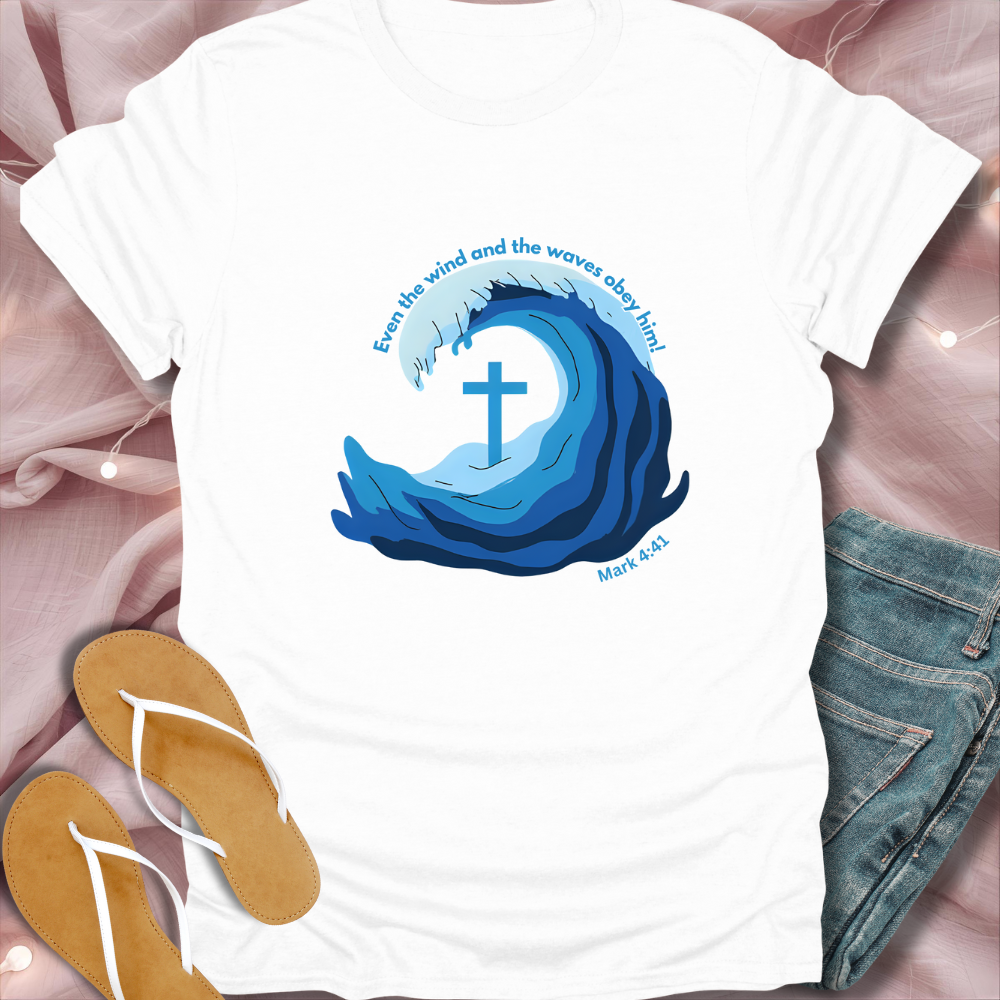 White t-shirt with blue wave design and cross, placed on a pink fabric background with sandals and jeans.