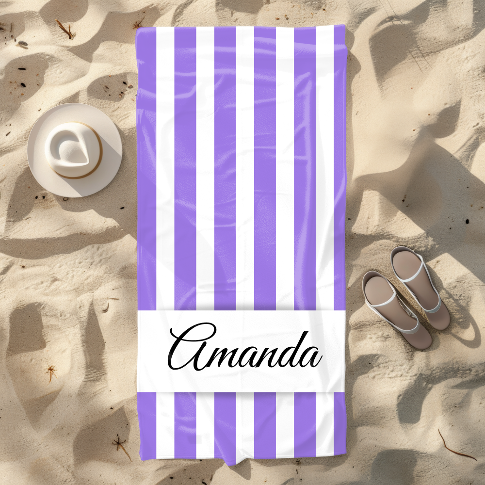 Beach towel with purple and white stripes and 'Amanda' branding on sand with hat and sandals.