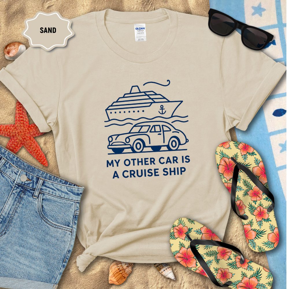 Beige t-shirt with a graphic design of a car and cruise ship, surrounded by beach items on sand.