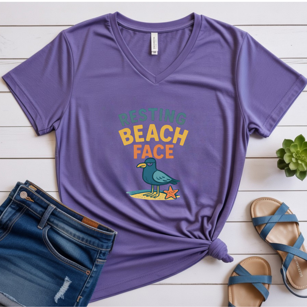 Purple t-shirt with 'Resting Beach Face' graphic on a wooden surface with jeans and sandals.
