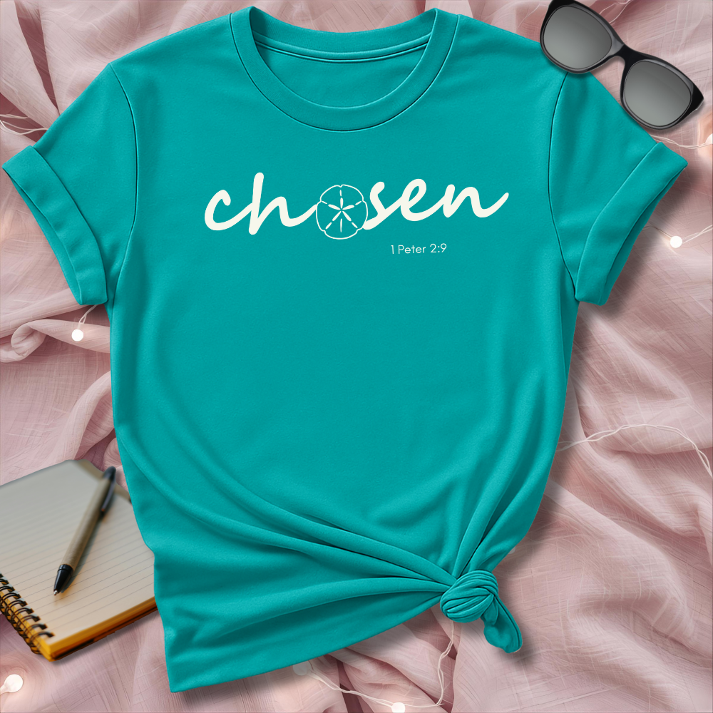 Turquoise t-shirt with 'chosen' text on a pink surface with sunglasses and notebook.