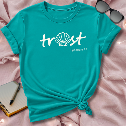 Turquoise t-shirt with 'trust' and 'Ephesians 1:7' text on a pink surface with sunglasses and notebook.