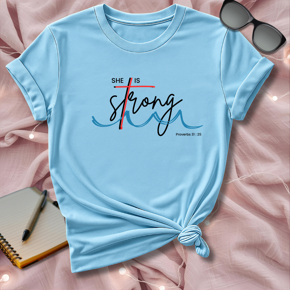 Light blue t-shirt with 'She is Strong' text on a pink surface with sunglasses and notebook.