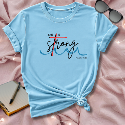 Light blue t-shirt with 'She is Strong' text on a pink surface with sunglasses and notebook.