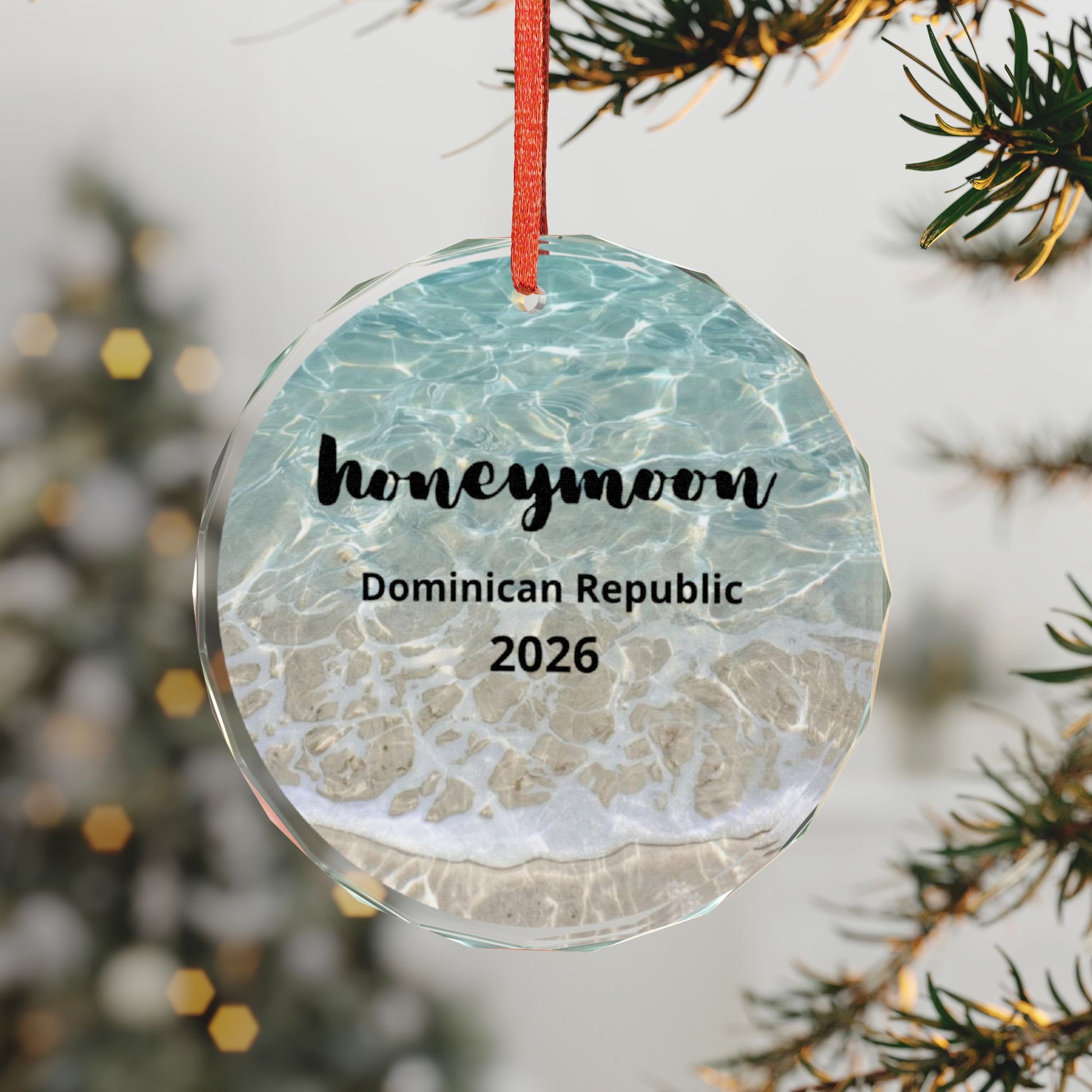 A round clear glass Christmas ornament with a red hanging ribbon, printed with a beach-and-sea pattern and the text 'honeymoon', 'Dominican Republic', and '2026'. The ornament is hanging in front of a blurred Christmas tree and pine branches.