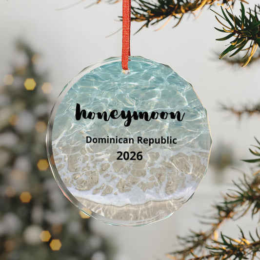 A round clear glass Christmas ornament with a red hanging ribbon, printed with a beach-and-sea pattern and the text 'honeymoon', 'Dominican Republic', and '2026'. The ornament is hanging in front of a blurred Christmas tree and pine branches.