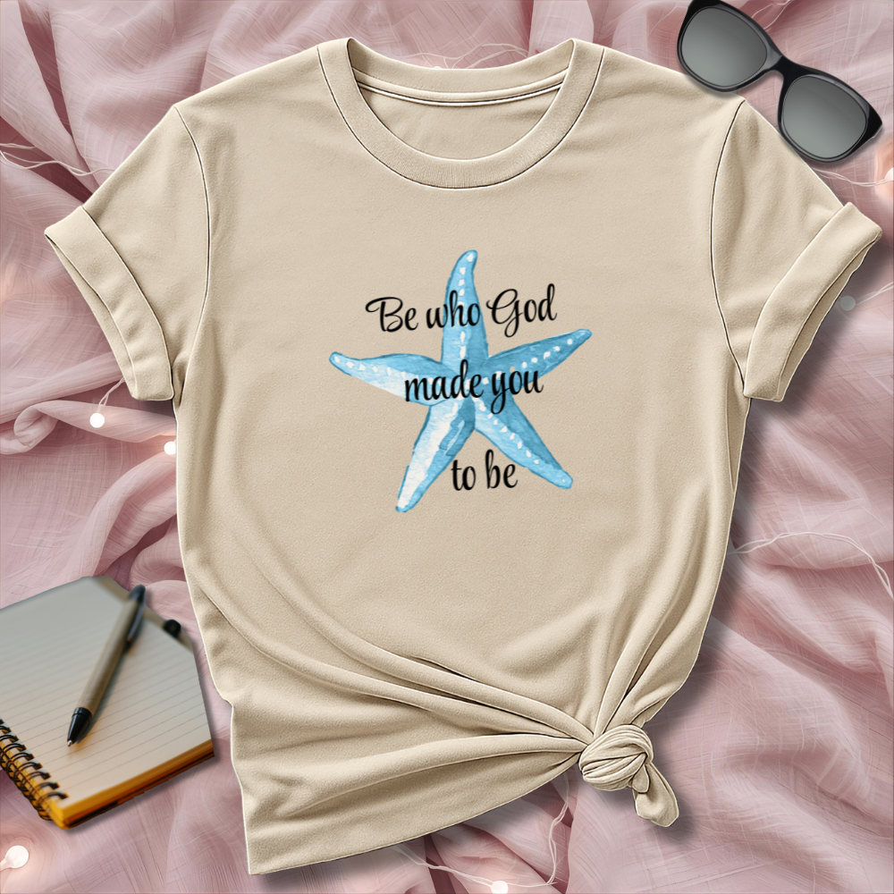 Beige t-shirt with blue starfish design and text on a pink surface with sunglasses and notebook.