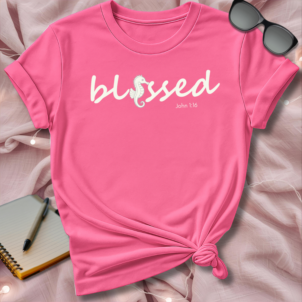 Pink t-shirt with 'blessed' text on a pink surface with sunglasses and notebook.