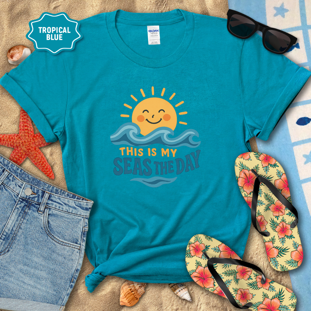 Blue t-shirt with sun and wave design, surrounded by beach accessories on sand.