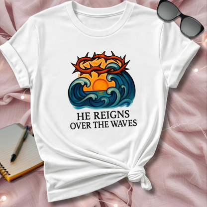 White t-shirt with a graphic design and text 'He Reigns Over the Waves' on a pink surface.