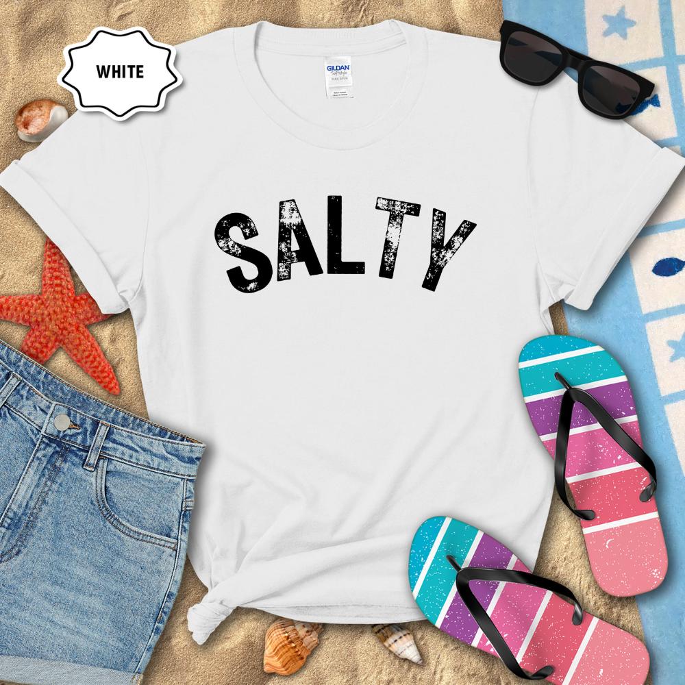 White t-shirt with 'SALTY' text on a sandy surface with beach accessories.