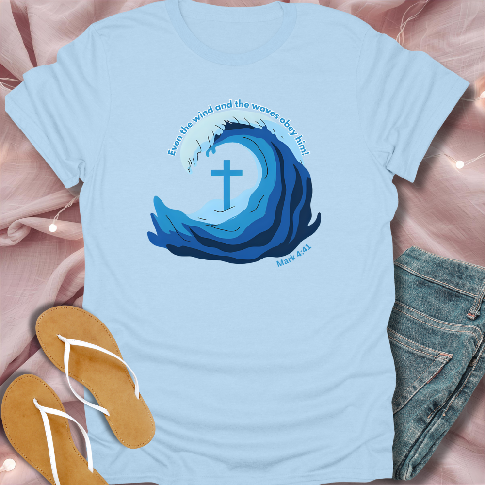Light blue t-shirt with wave design and cross, placed on a pink fabric background with sandals and jeans.
