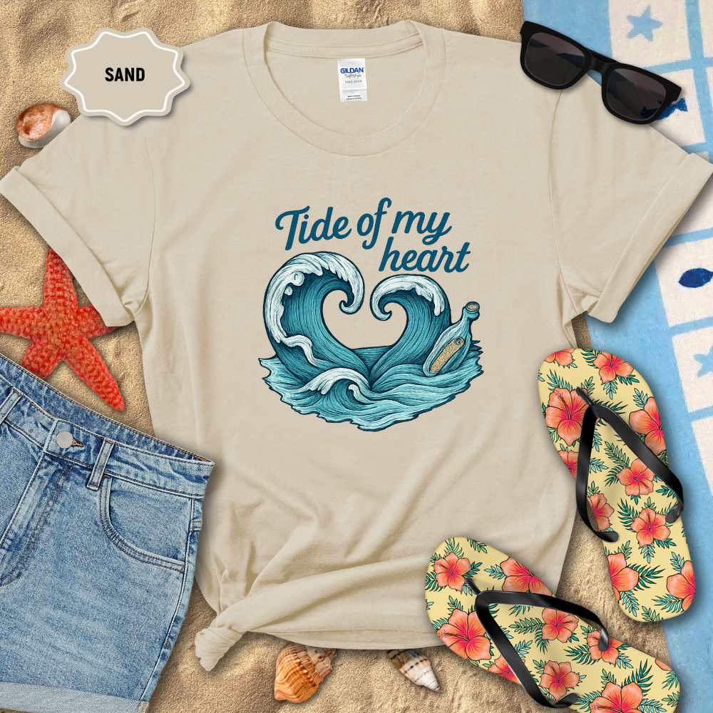 Beige t-shirt with wave design and text 'Tide of my heart' on a sandy background with beach accessories.