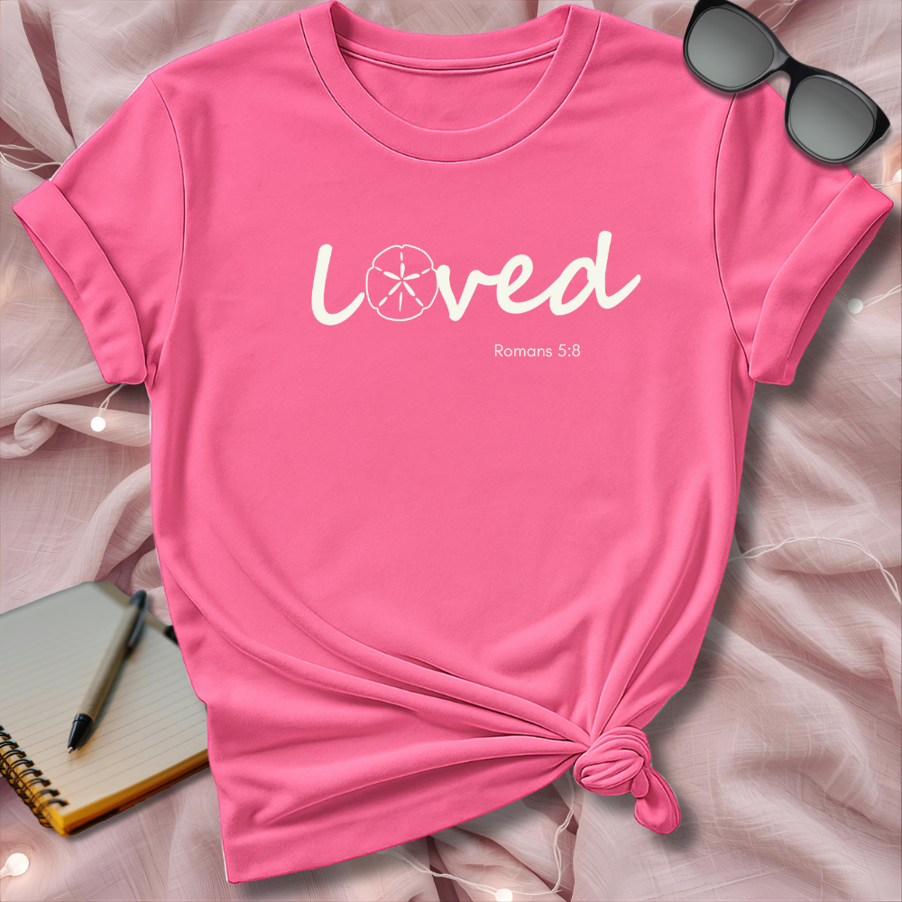 Pink t-shirt with 'Loved' text on a soft pink surface with sunglasses and notebook.