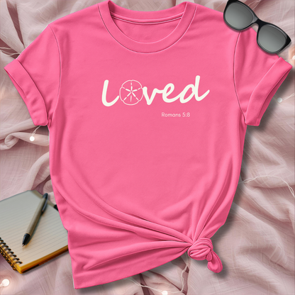 Pink t-shirt with 'Loved' text on a soft pink surface with sunglasses and notebook.
