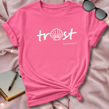 Pink t-shirt with 'trust' and shell design on a pink surface with sunglasses and notebook.