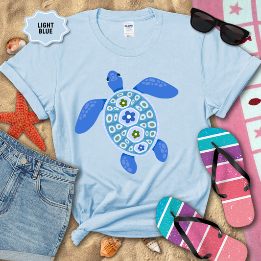 Light blue t-shirt with a turtle design on a sandy background with beach accessories.