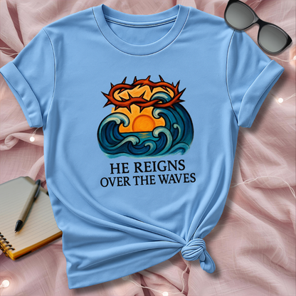 Light blue t-shirt with a graphic design and text 'He Reigns Over The Waves' on a pink surface.