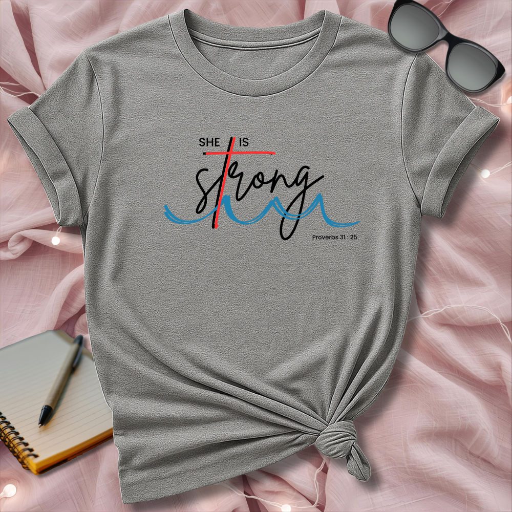 Gray t-shirt with 'She is Strong' design on a pink surface with sunglasses and notebook.