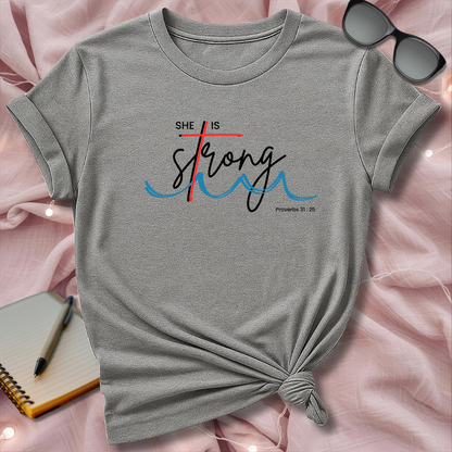 Gray t-shirt with 'She is Strong' design on a pink surface with sunglasses and notebook.