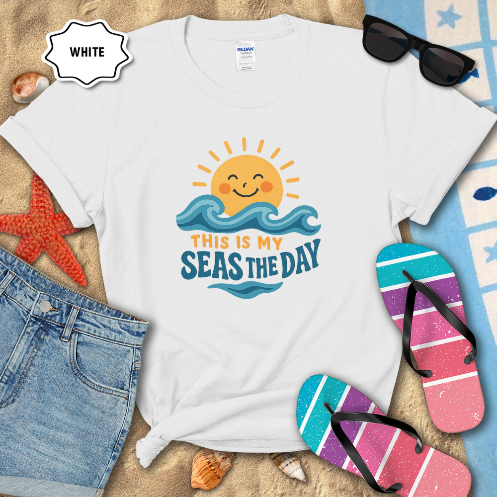 White t-shirt with beach-themed design on sand with sunglasses, flip-flops, and denim shorts.
