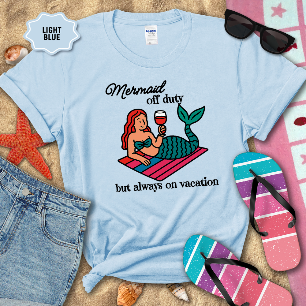 Light blue t-shirt with mermaid graphic and text on a sandy background with beach accessories.