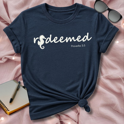 Navy blue t-shirt with 'redeemed' design on a pink surface with sunglasses and notebook.
