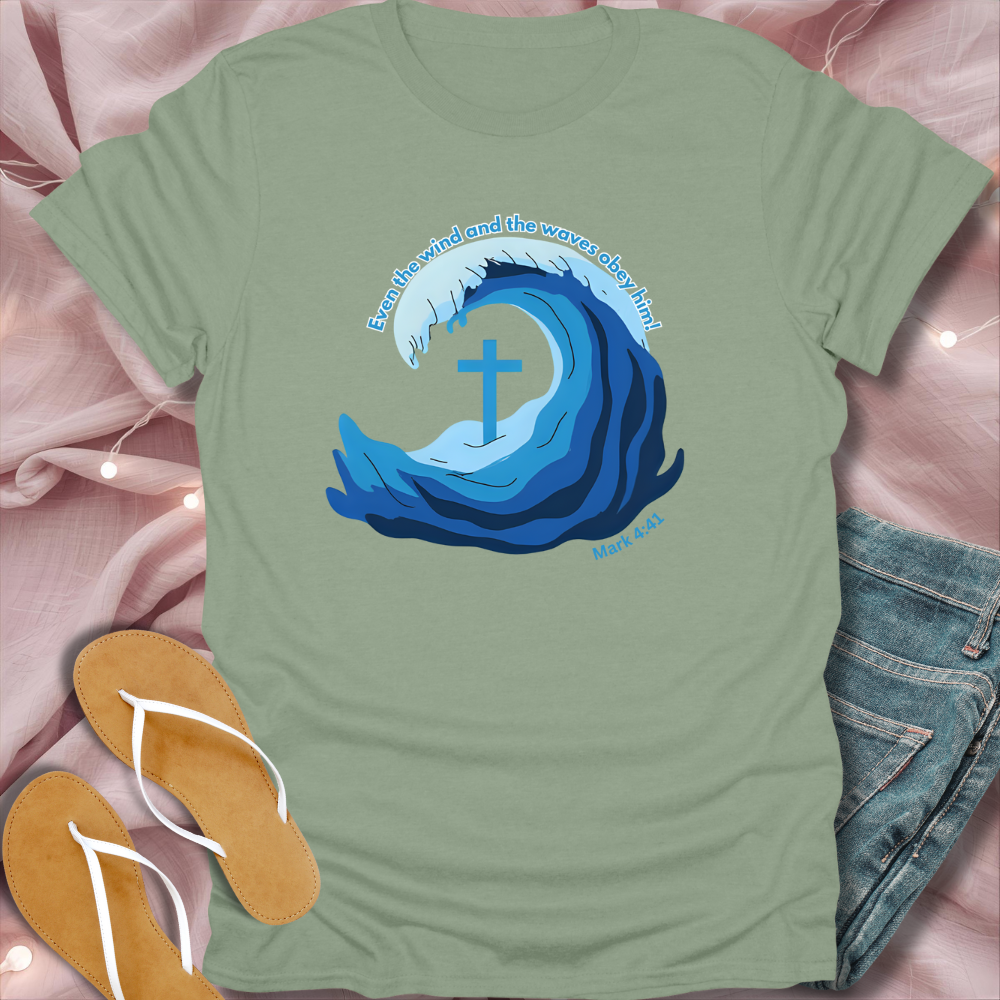 Green t-shirt with blue wave design and cross, placed on a pink fabric background with sandals and jeans.