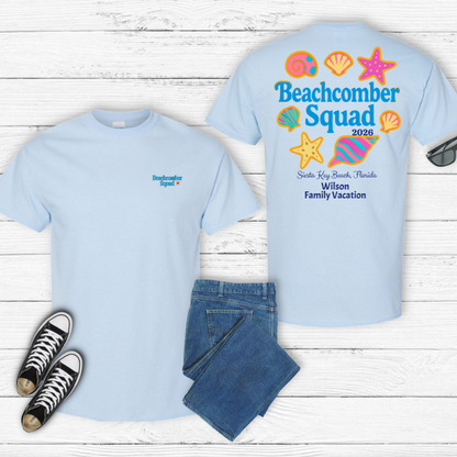 Two light blue t-shirts with 'Beachcomber Squad' design on a wooden surface, with jeans and sneakers.