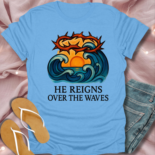 Blue t-shirt with a graphic design and text 'He Reigns Over the Waves' on a pink fabric background.
