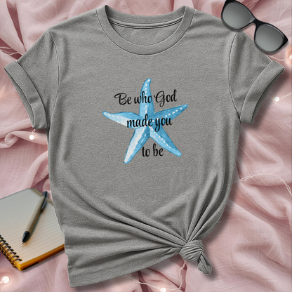 Gray t-shirt with blue starfish design and text on a pink surface with sunglasses and notebook.