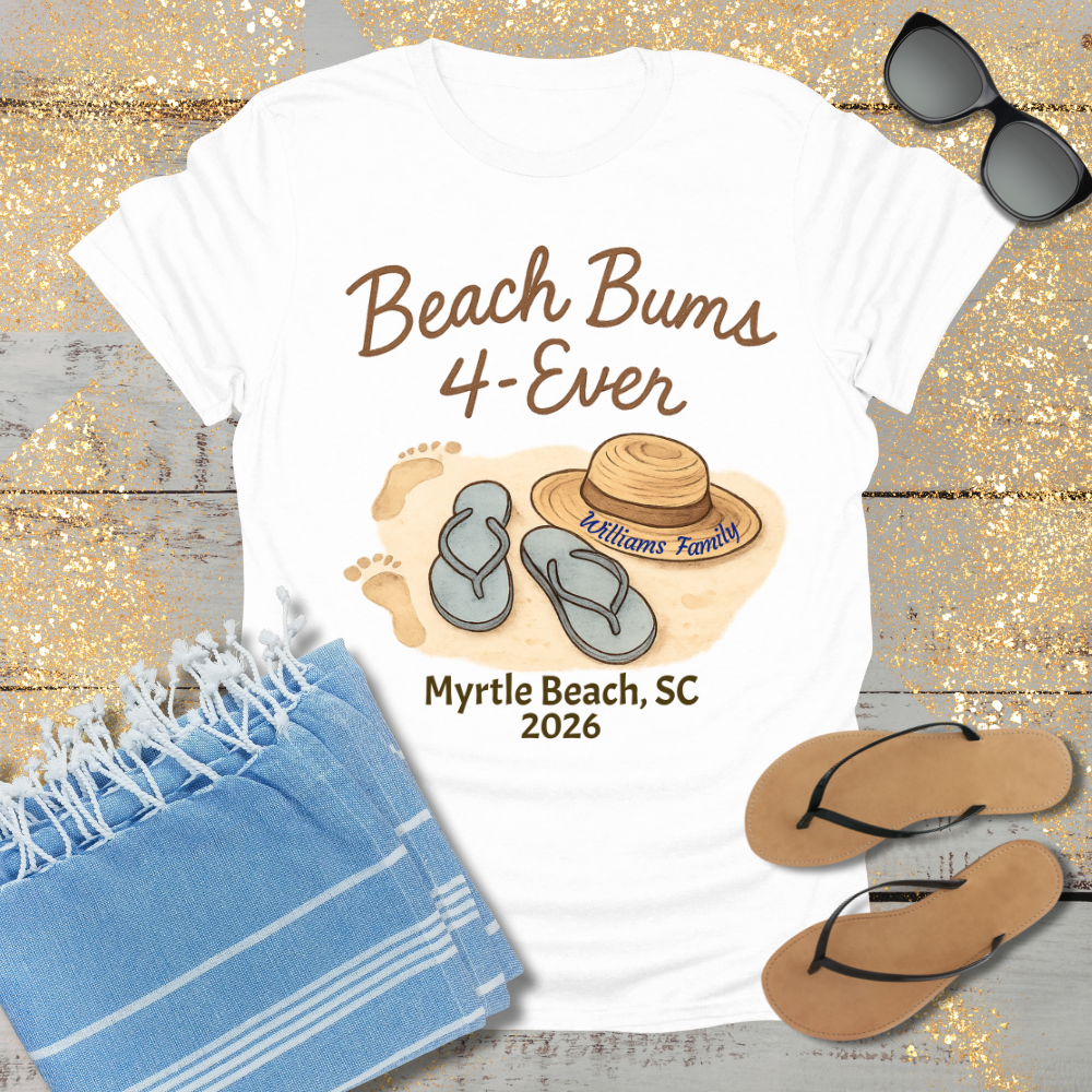 White t-shirt with 'Beach Bums 4-Ever' design featuring flip-flops and a hat, placed on a wooden surface with sunglasses and a towel.