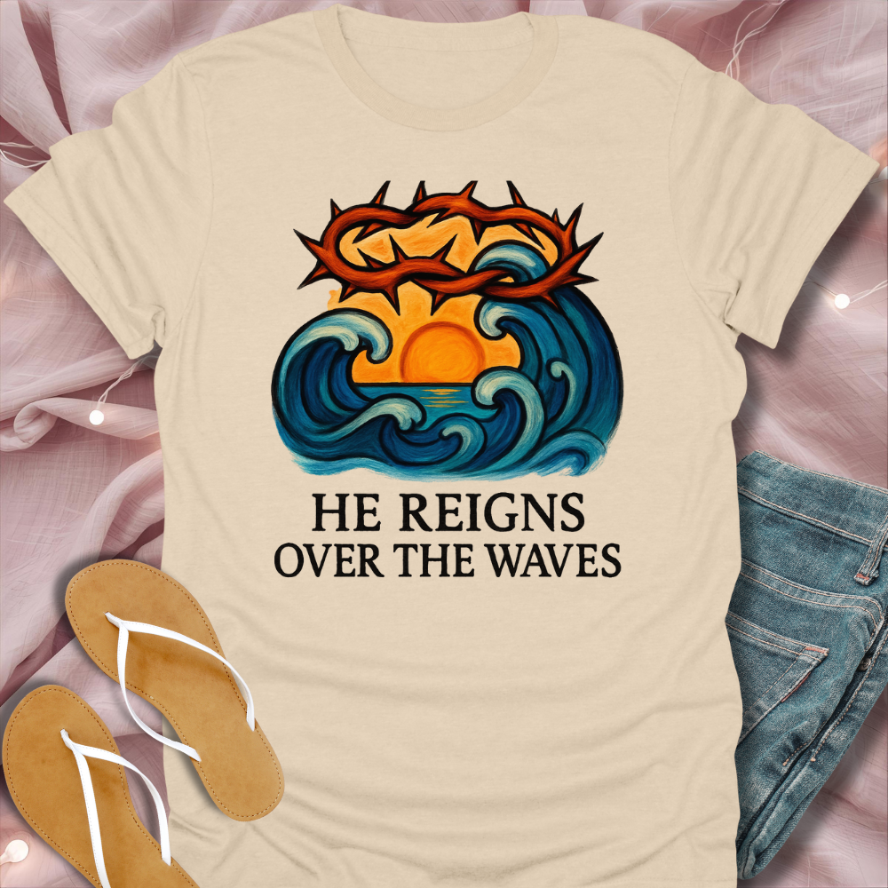 Beige t-shirt with a graphic design of waves and a crown with text 'He Reigns Over the Waves' on a pink surface.