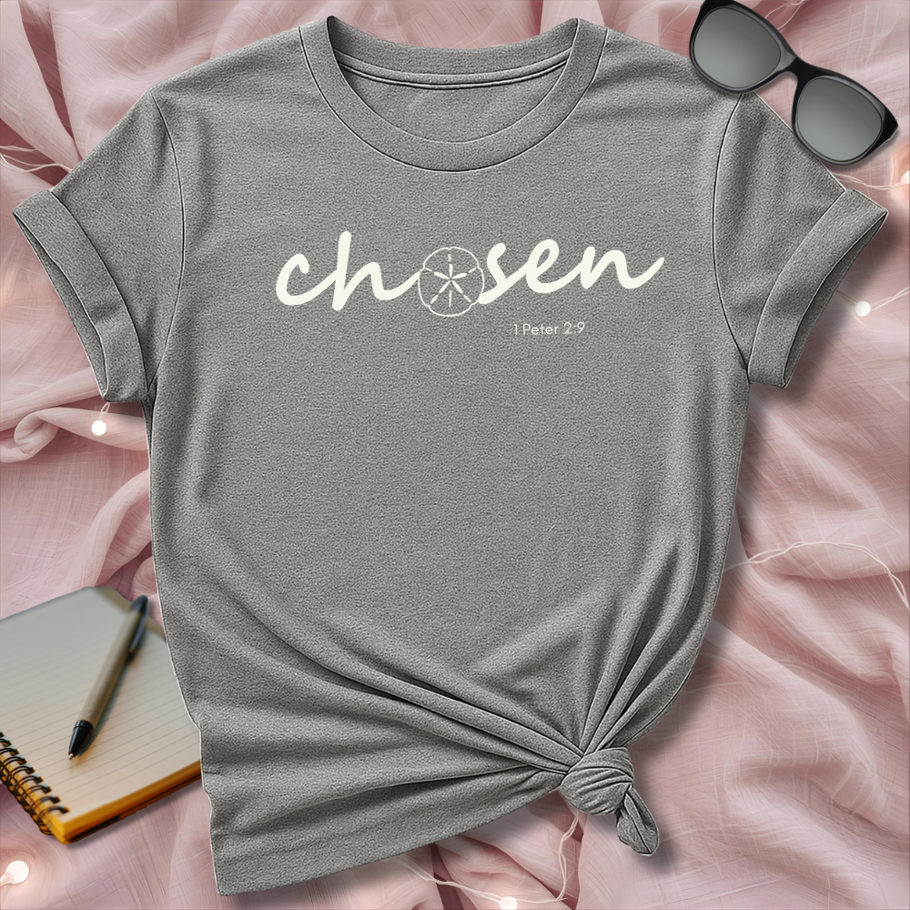 Gray t-shirt with 'chosen' text on a pink surface with sunglasses and notebook.